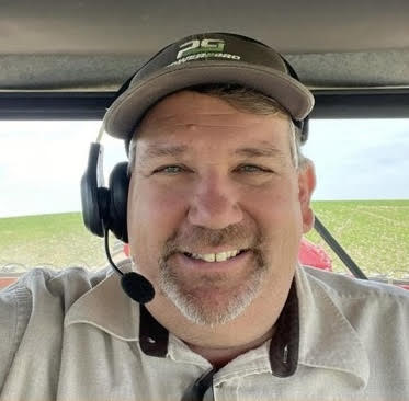 Selfie of a man smiling while wearing a baseball hat and headset taken inside the cab of a tractor