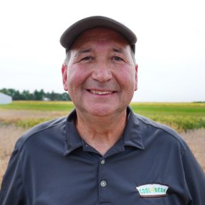 A man in  a cap and blue shirt smiles in front of a field of crops